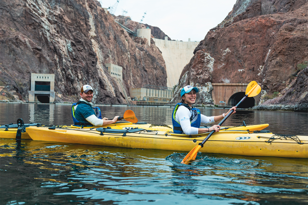 Two people kayaking on a river near a dam, surrounded by rocky cliffs.