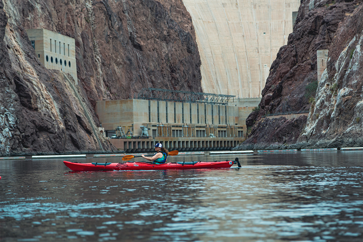 Person kayaking on a river beneath a large dam and rocky cliffs.
