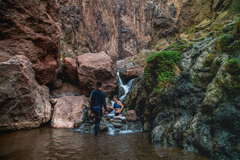 Two people near cascading waterfall in a rocky canyon setting.