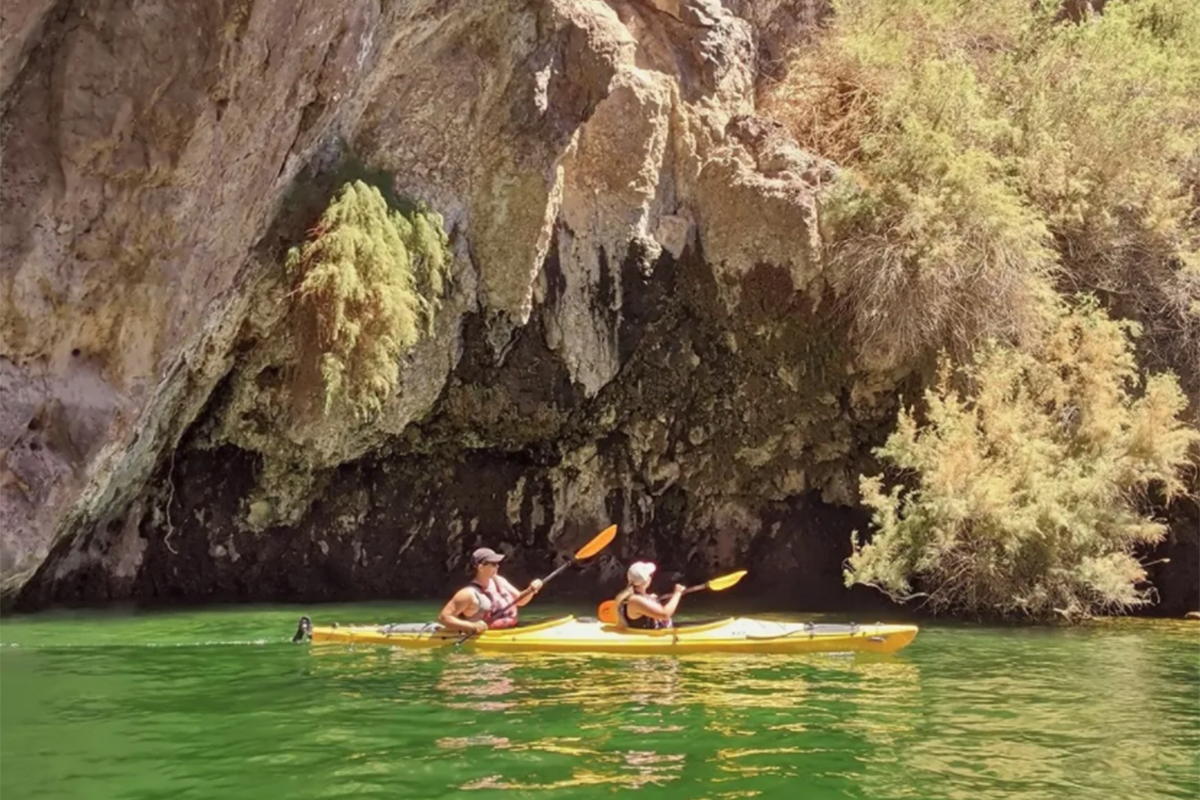 Two people kayaking on a green river, near rocky cliffs and trees.