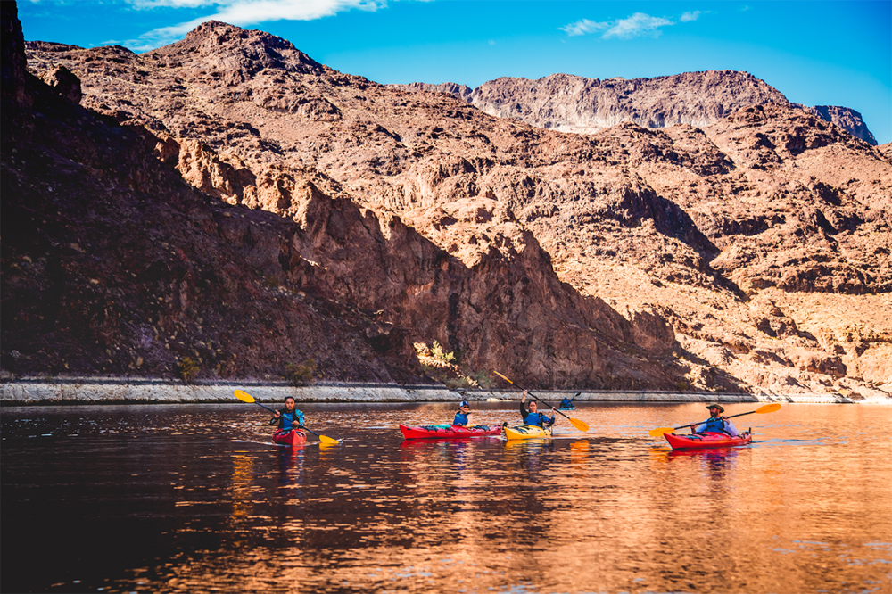Four people kayaking on a calm river with rocky hills in the background.