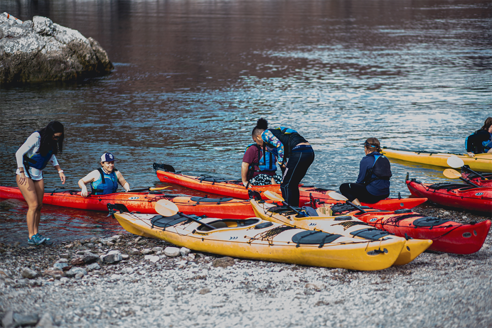 People in life jackets prepare kayaks on a rocky shore before kayaking.