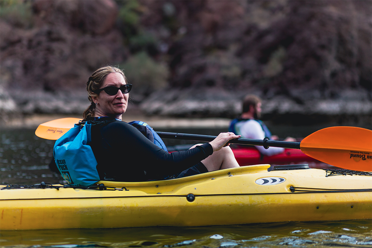 Woman in sunglasses kayaking on a river with another kayaker in the background.