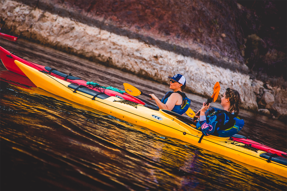 Two people kayaking in a yellow kayak on a calm lake near rocky shore.