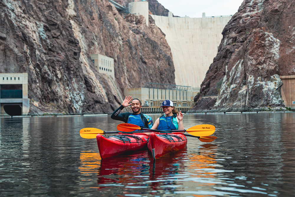 Two people kayaking on a river near a large dam with rocky cliffs.