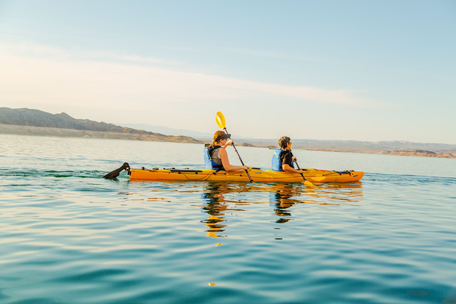 Two people kayaking on a calm lake with mountains in the background.