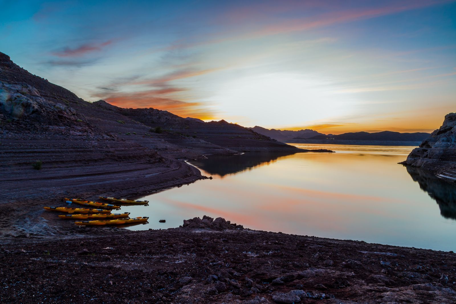 Sunrise over a calm lake with yellow kayaks on a rocky shore and silhouetted hills.