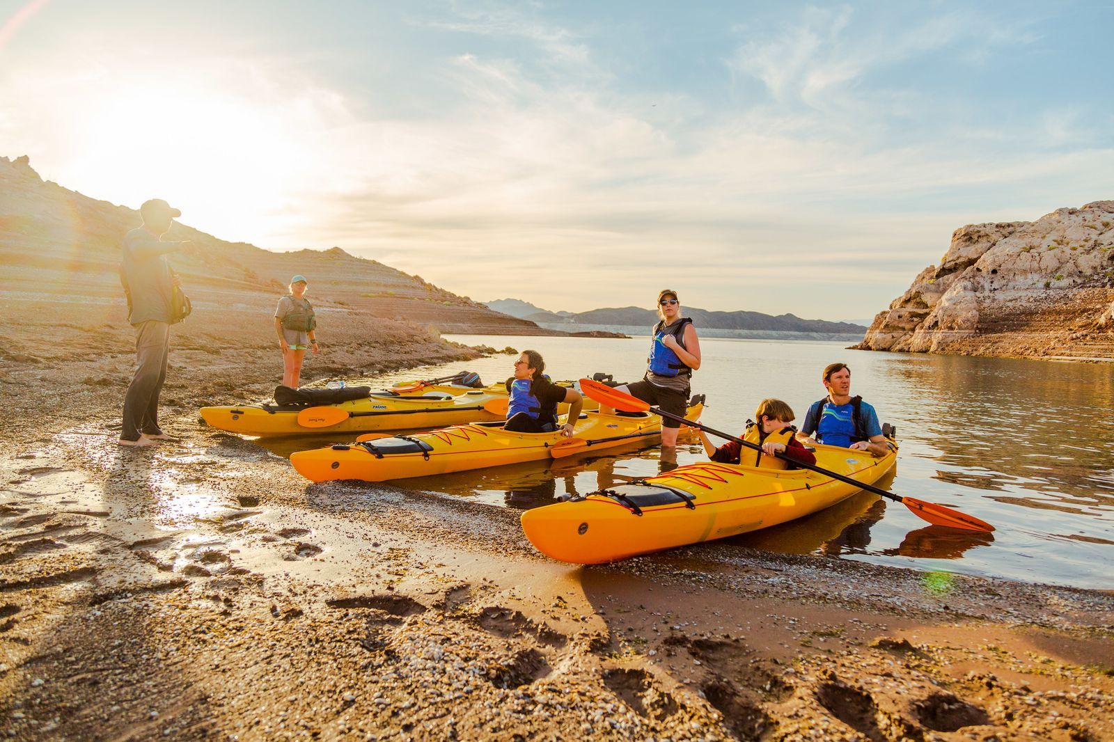 Group of people with kayaks on a beach by a lake at sunset.