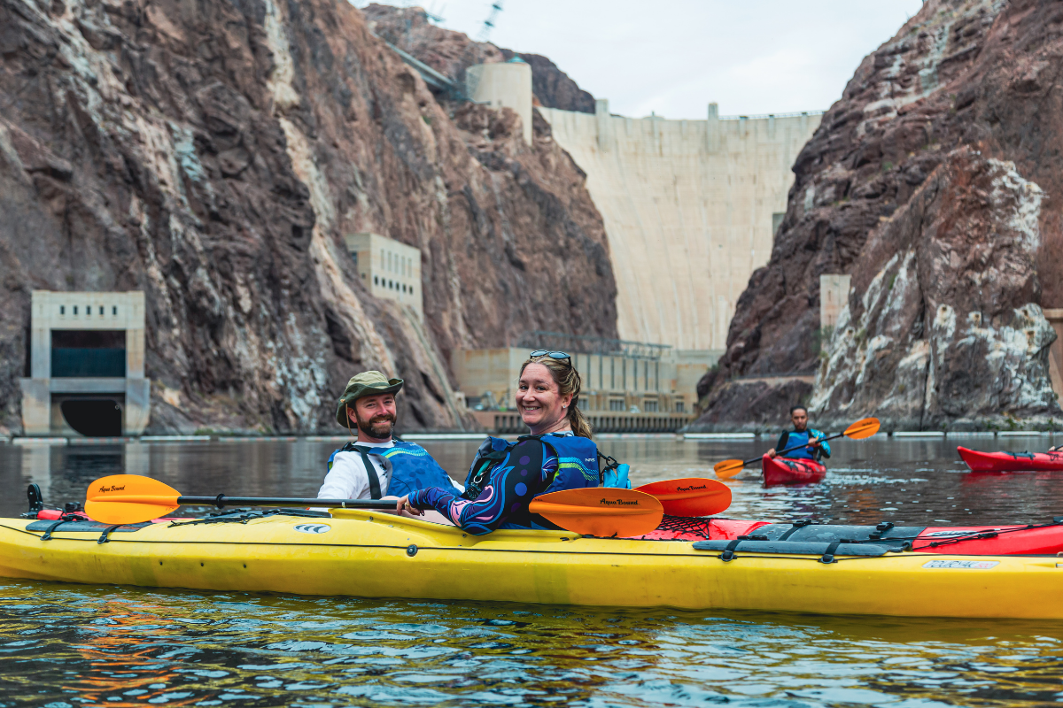 People kayaking on a river near a large dam and rocky cliffs.
