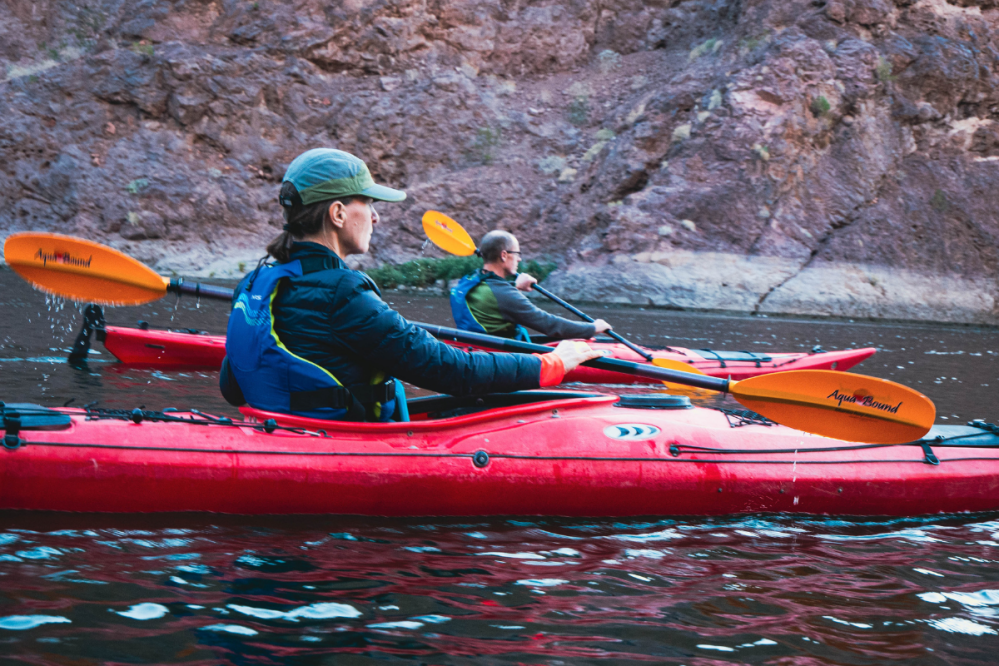 Two people kayaking in red kayaks on a river with rocky cliffs in the background.