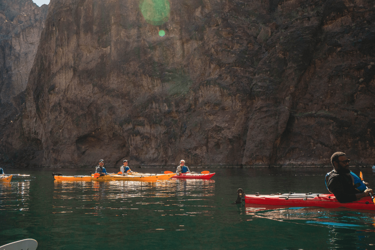 Group kayaking on a calm river by steep rock cliffs, under sunlight.