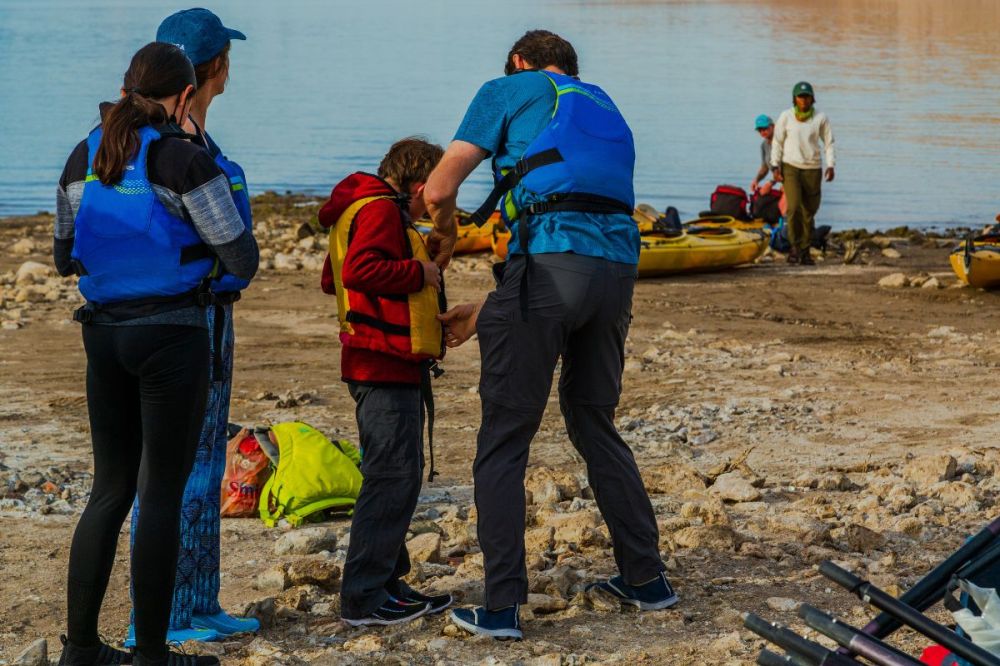 People in life jackets preparing kayaks by a lake shore.