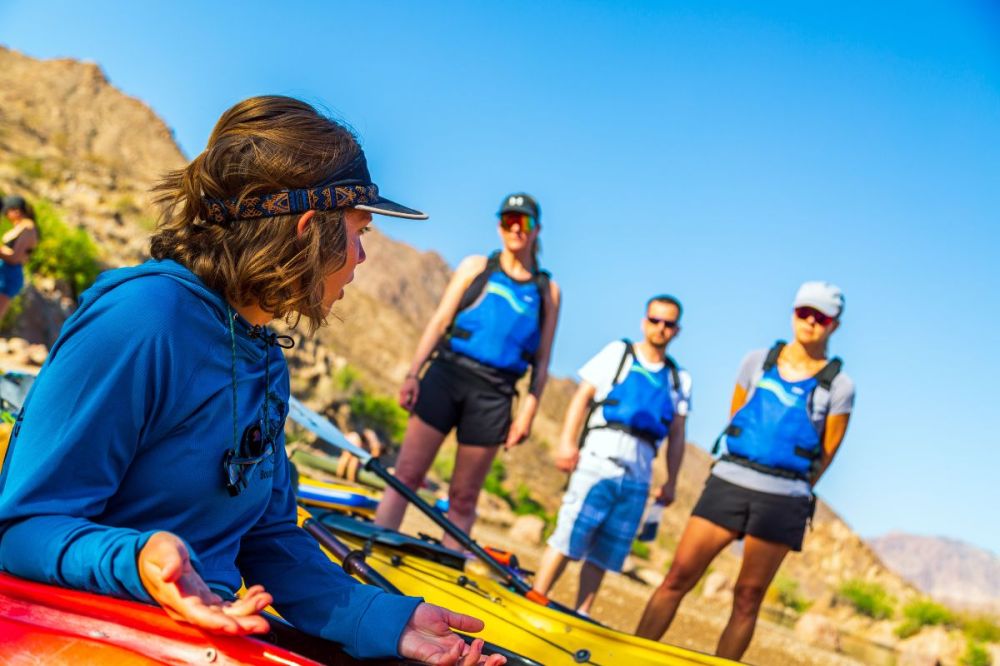 Person in blue talking to a group in life vests near kayaks on a sunny day.