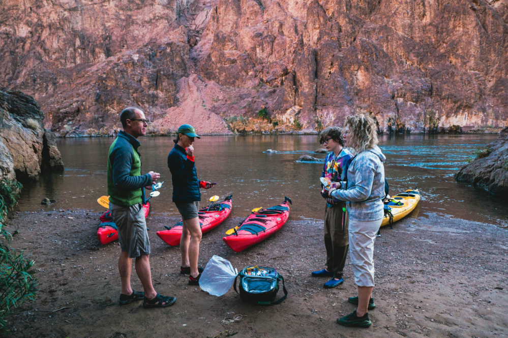 Four people stand by kayaks on a riverbank with rocky cliffs in the background.