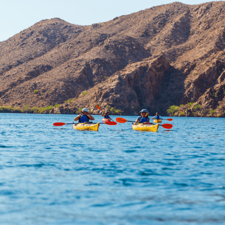 Two kayakers paddle on a lake with large rocky hills in the background.