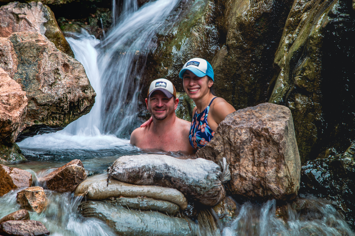 Two people wearing caps smiling in a rocky waterfall pool.