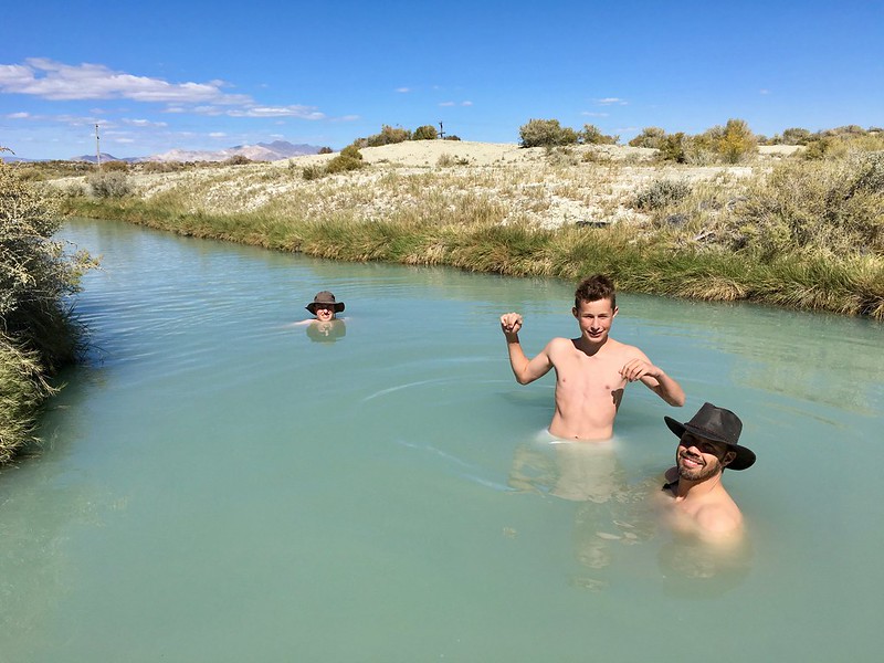 Three people wearing hats are wading in a shallow, blue water stream on a sunny day.