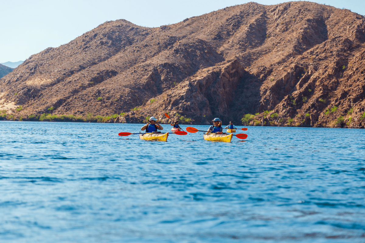 Two people kayaking on a blue lake with rocky hills in the background.