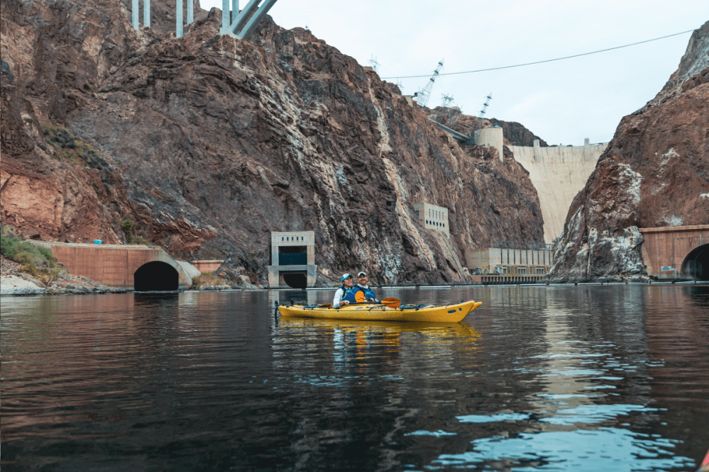 Two people kayaking on a river near a large dam and rocky cliffs.