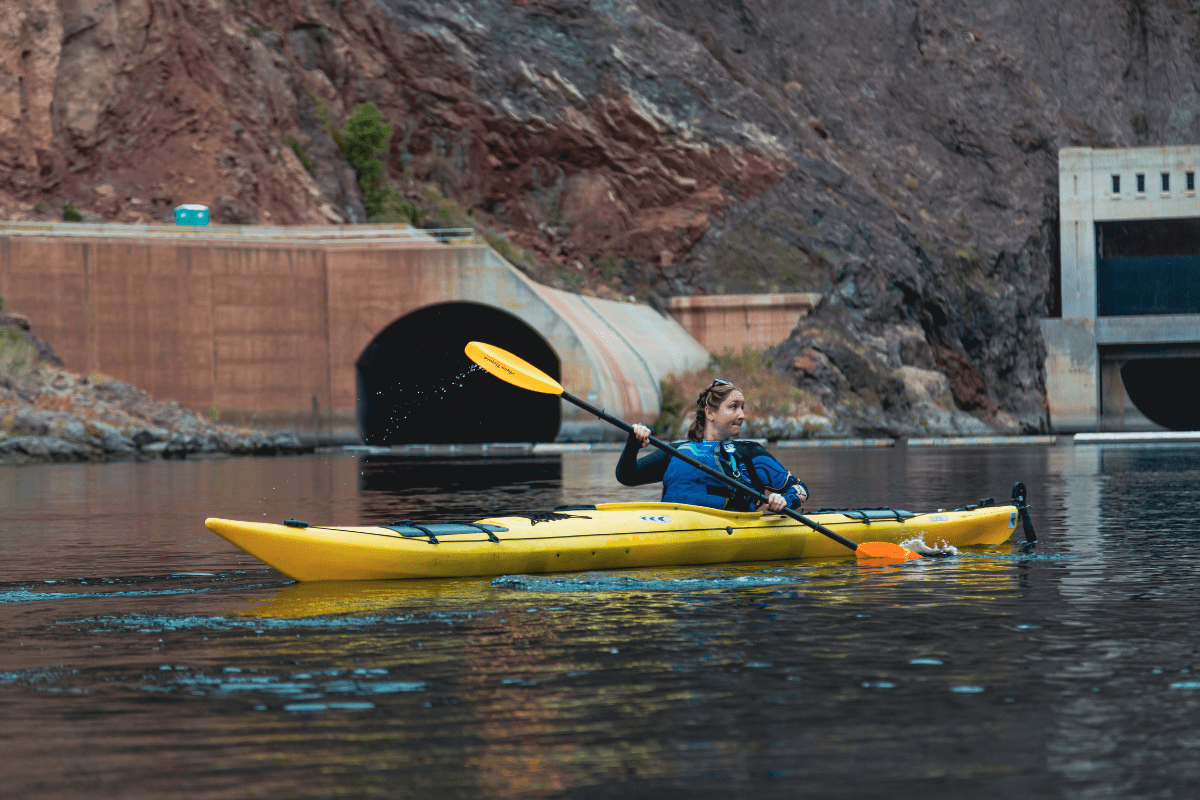 Person kayaking in a yellow kayak on a calm river near a tunnel and rocky cliffs.