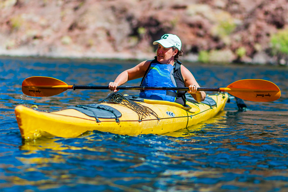 Person kayaking on a lake with a yellow kayak and wearing a blue life vest and cap.