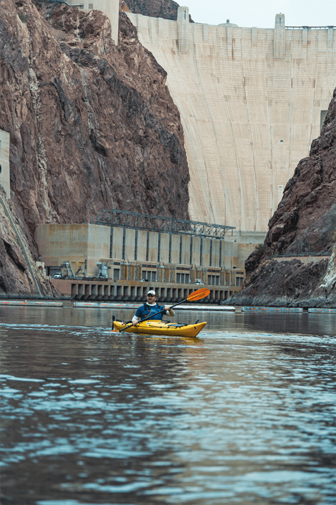 Person kayaking on a river in front of a large concrete dam with rocky cliffs.