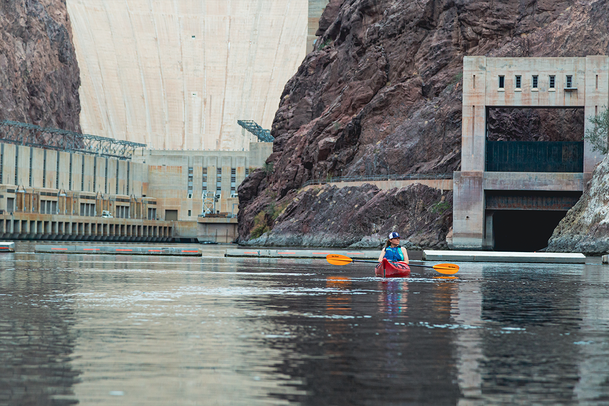 Person kayaking on a river near a large dam and rocky cliffs.