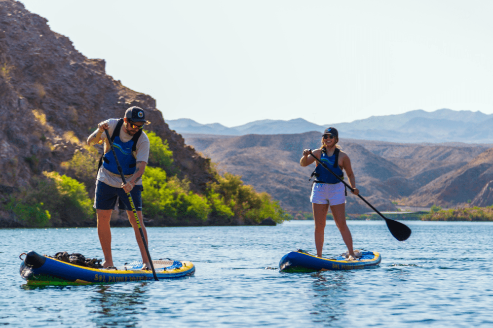Two people paddle boarding on a river with mountains in the background.