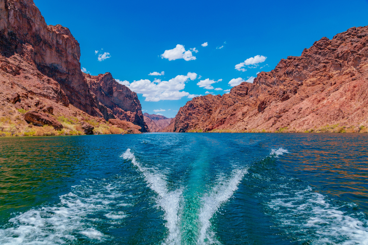 Boat wake on blue river between red rocky canyon walls under a bright blue sky.