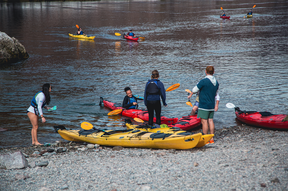 People with kayaks by a river, some launching, others paddling in the water.