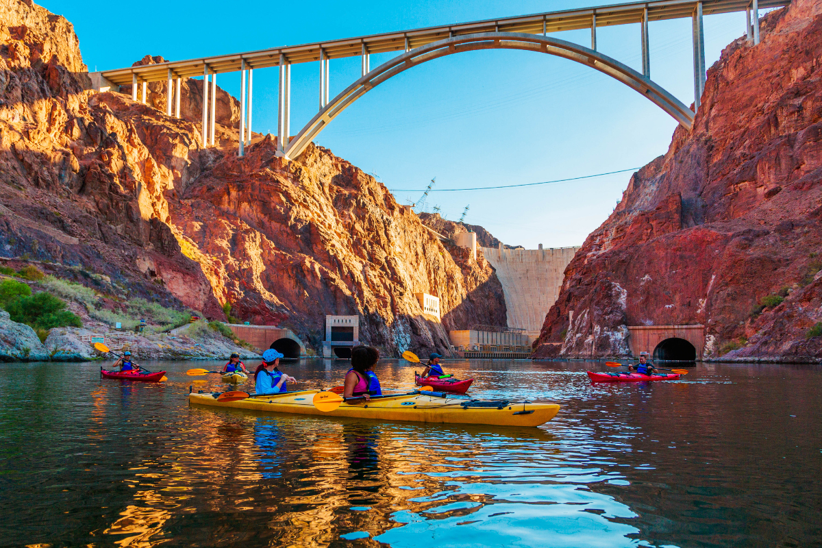 People kayaking on a river under a large bridge and between rocky cliffs.