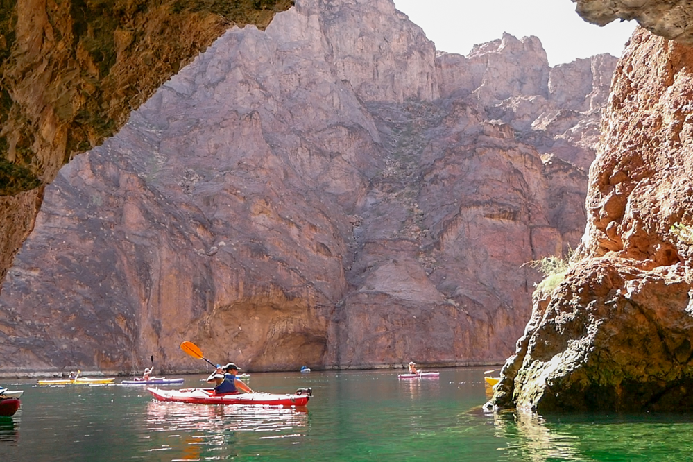 Kayakers paddle through a rocky canyon with steep cliffs and sunlight reflecting on the water.