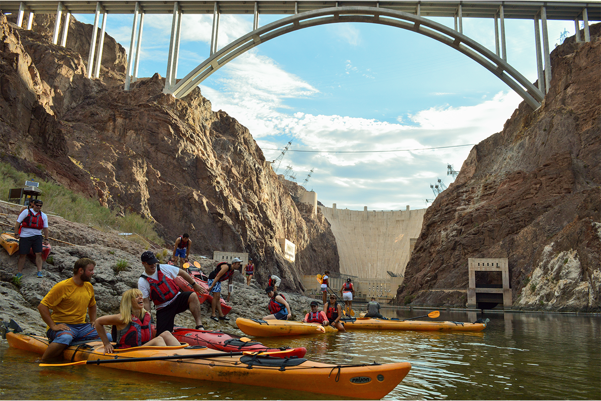 Group kayaking under a large arch bridge with rocky cliffs and dam background.