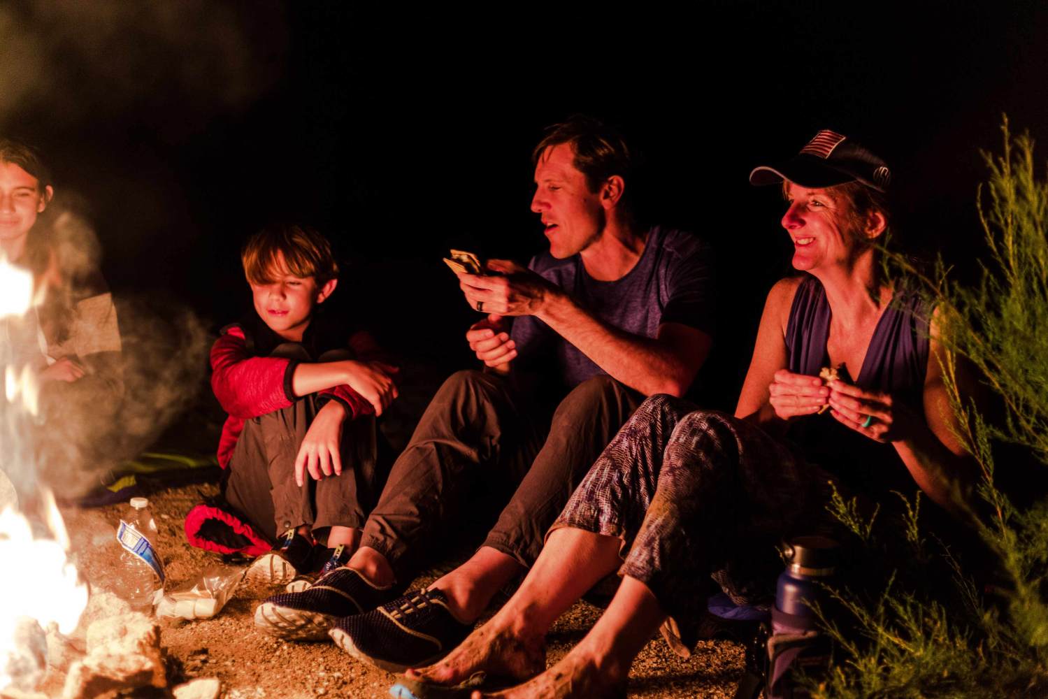 People sitting by a campfire at night, eating and smiling.