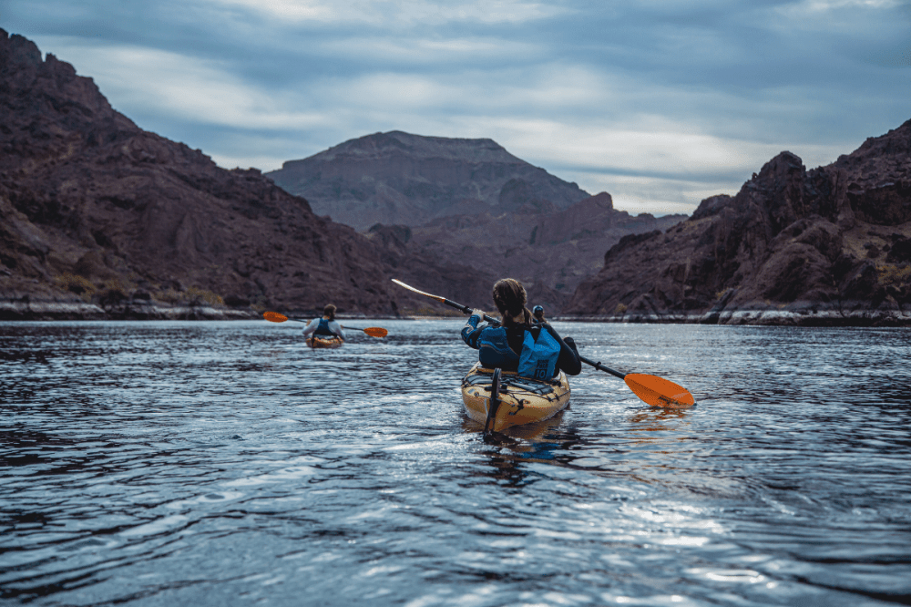 Two kayakers paddle on a calm river surrounded by rocky mountains under a cloudy sky.