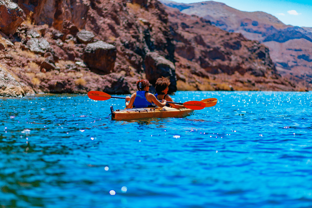 Two people kayaking on a blue lake with rocky hills in the background under a clear sky.