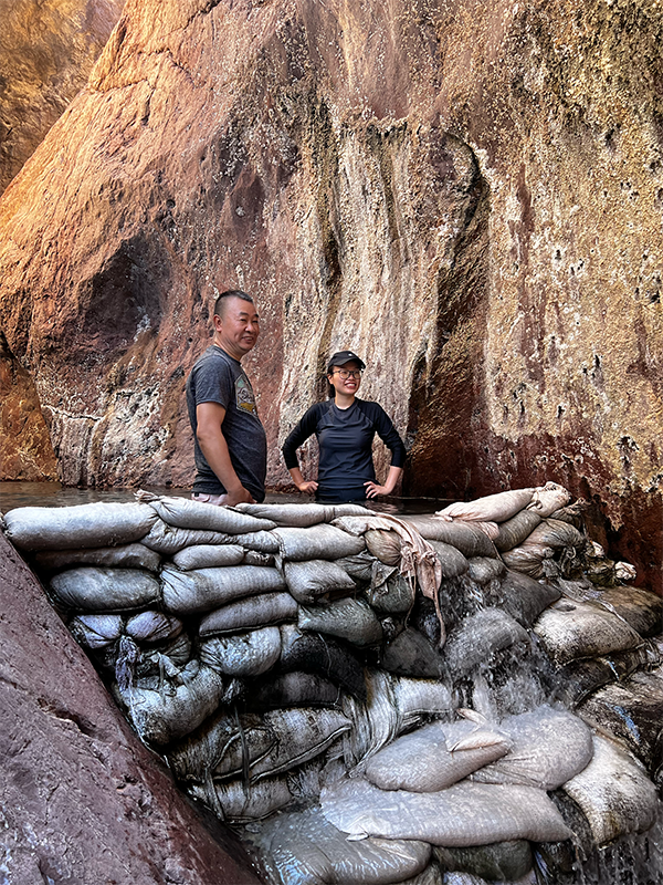 Two people standing behind sandbags with flowing water in a rocky canyon.