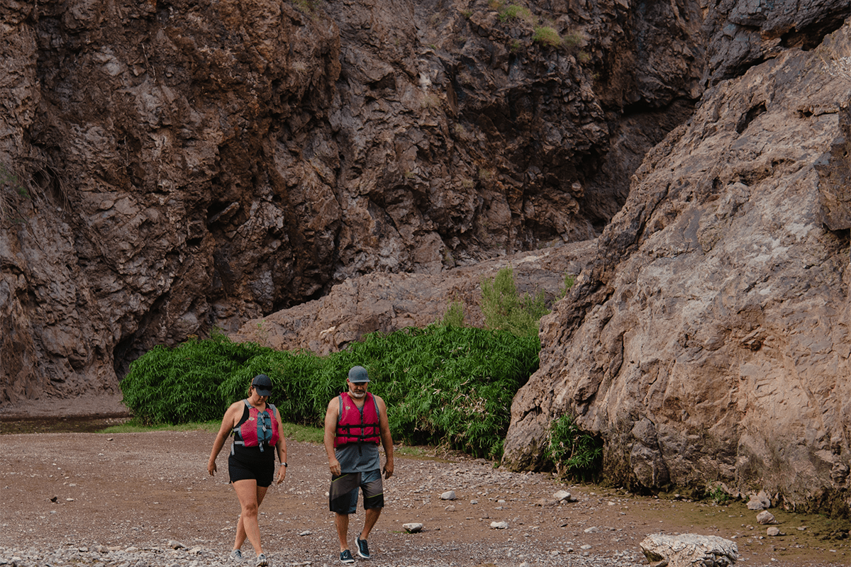 Two people wearing life jackets walk near rocky cliffs and greenery.
