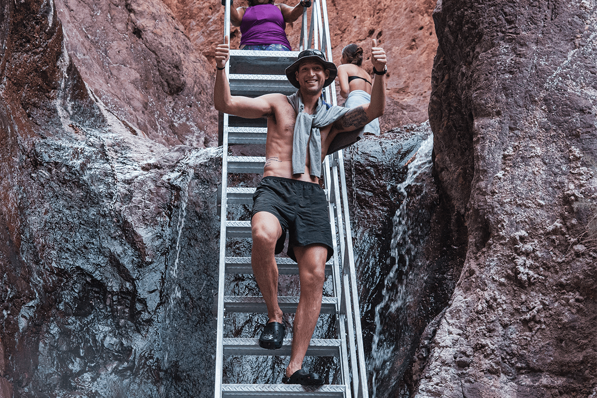 Person in shorts giving thumbs up on metal ladder in rocky canyon with small waterfall.