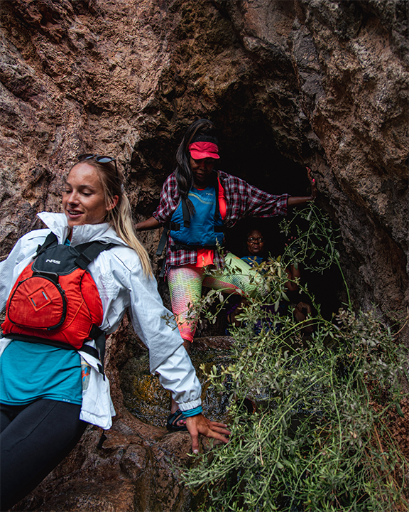 Three hikers navigate a rocky cave entrance with plants nearby.