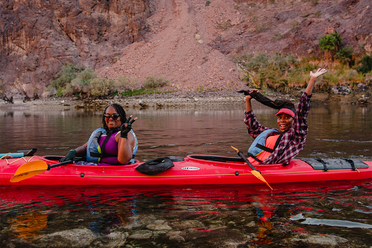 Two people in a red kayak smiling and gesturing on a calm river with rocky cliffs in the background.