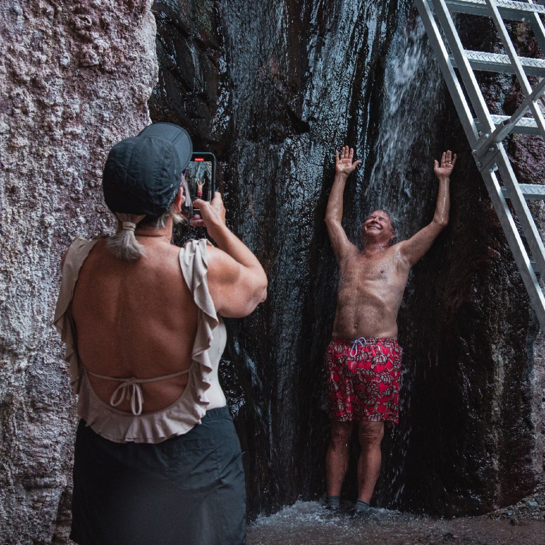 Woman photographs man posing under waterfall near rocky surface and ladder.