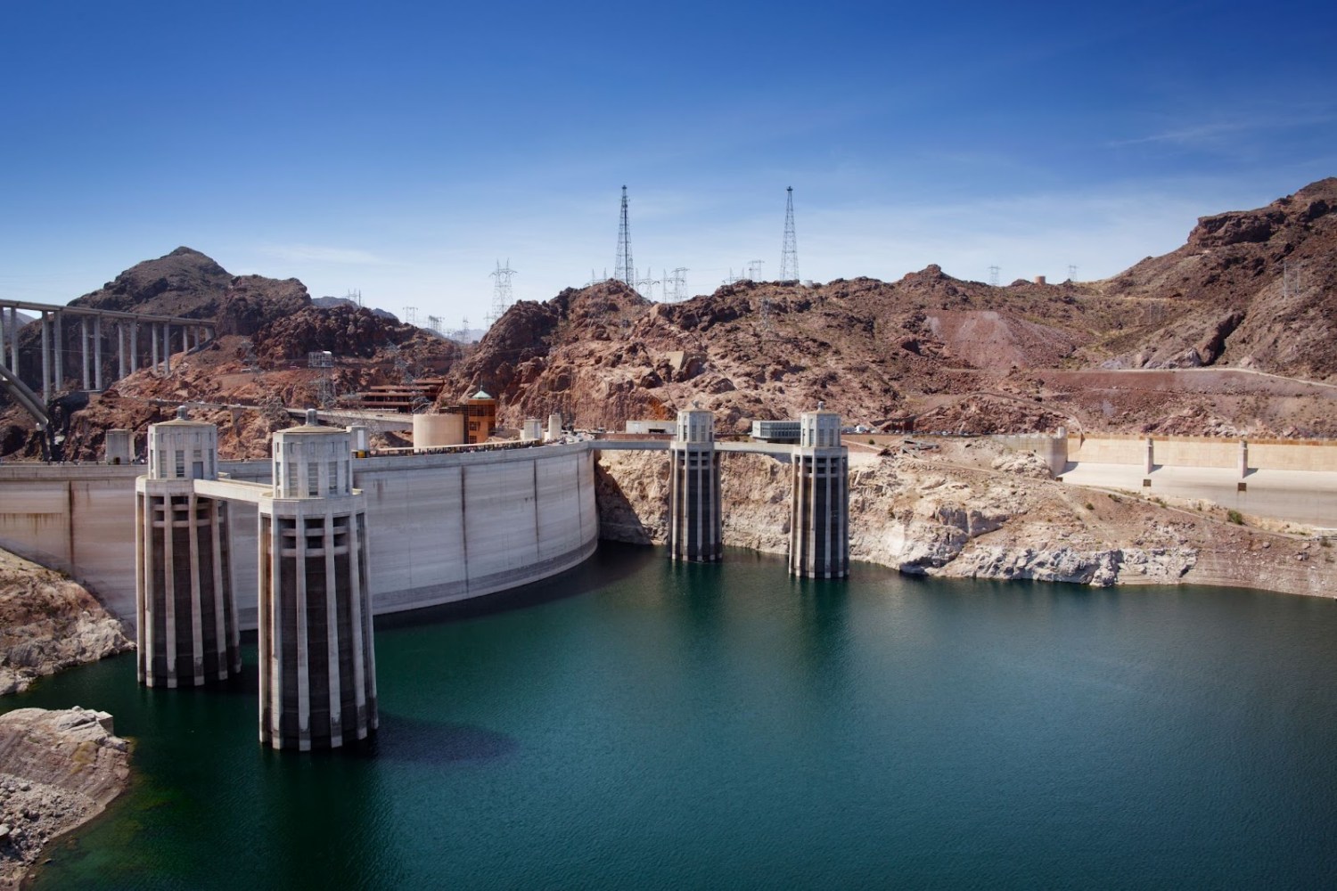 Concrete dam with towers in rocky landscape under a clear blue sky.
