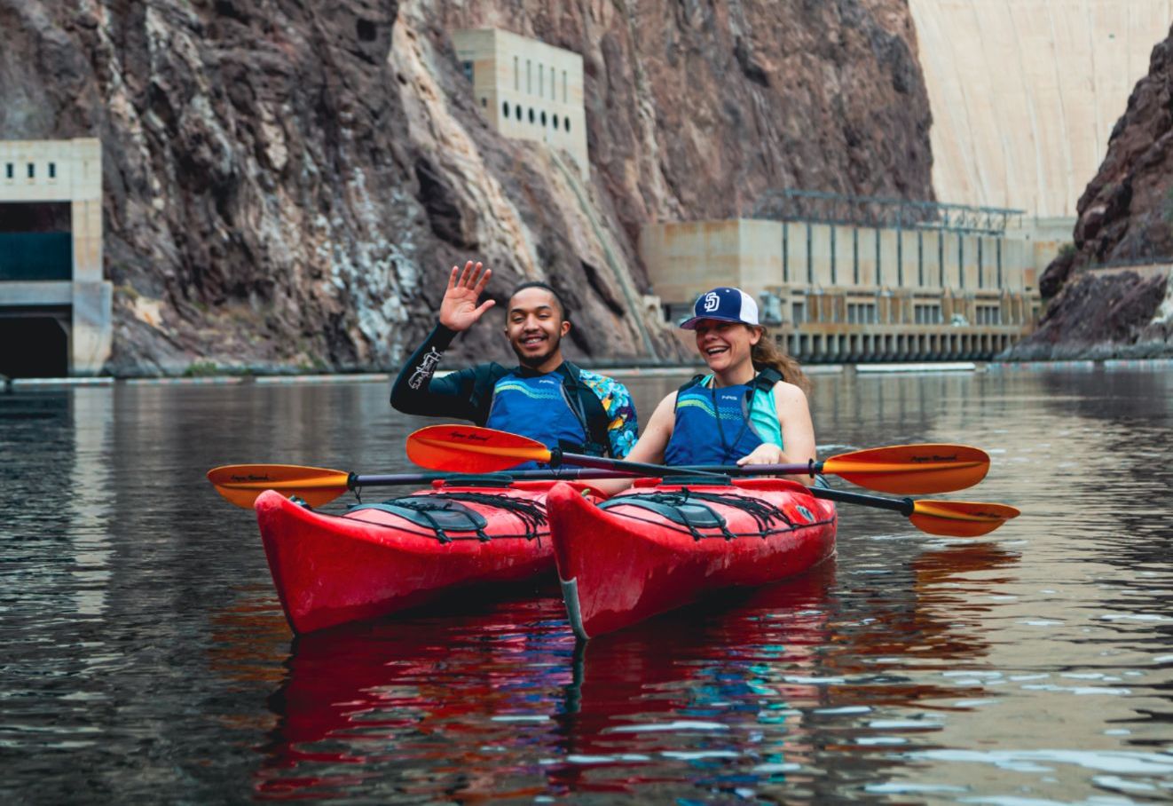 Two people in red kayaks smiling on a lake near rocky cliffs and a large dam.