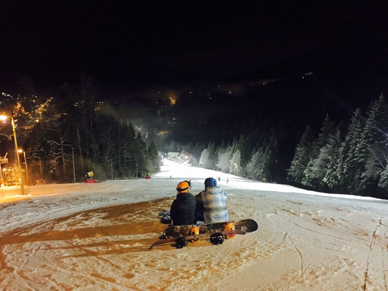 Two snowboarders sitting on a snowy slope at night, facing a lit village.