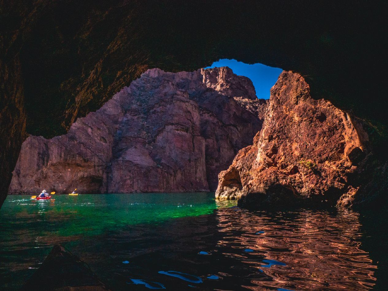 Kayakers on a green lake in a cave with rocky walls and a bright blue sky.