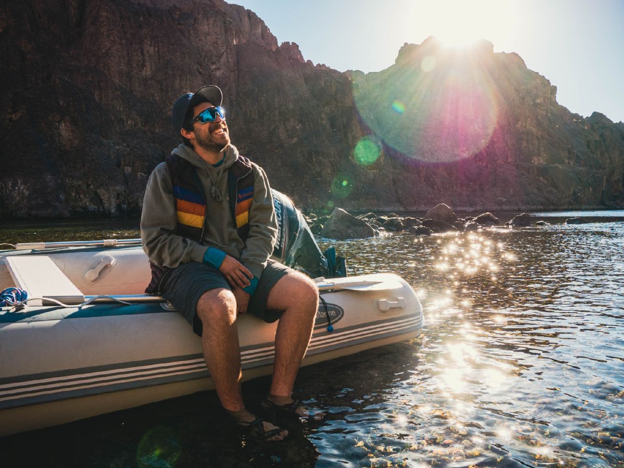Man sitting on an inflatable boat, smiling, with sunlit mountains and water in the background.