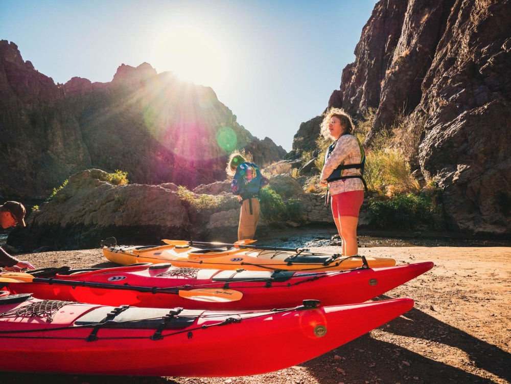 People near colorful kayaks on a sandy bank with a sunlit rocky background.
