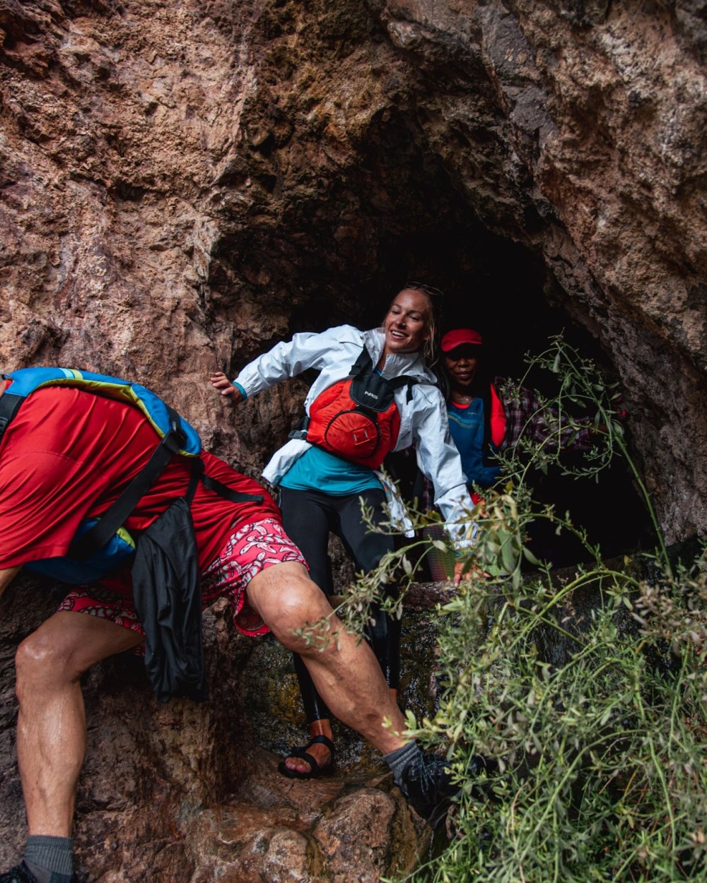 Hikers navigating rocky terrain and foliage, one wearing a red helmet and another in a white jacket.