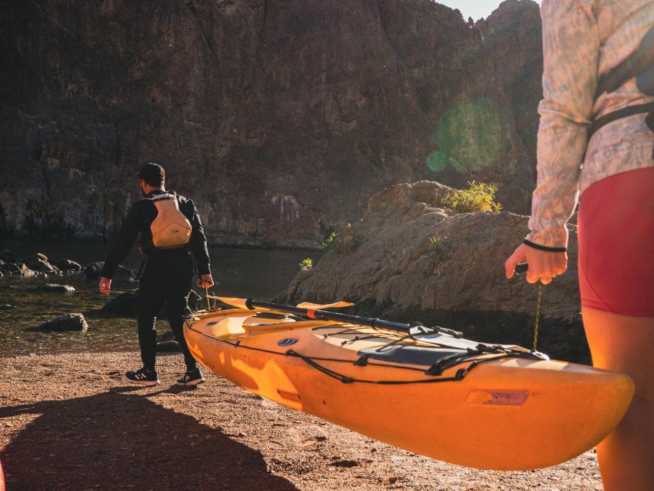 Two people carrying a yellow kayak towards a river, surrounded by rocky terrain.